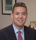 Anthony Shih has short gray hair and is wearing a suit. There are several frames on the wall behind him.