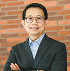 Gilbert Gee has short black hair and glasses. He is standing in front of a brick wall with his arms crossed.
