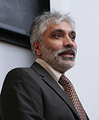Sanjay Gulati has gray hair and facial hair. He is wearing a brown suit and looking off to the side.