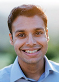 Joshua Budhu has short black hair. He is standing outside and wearing a blue shirt.