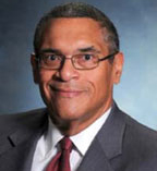 Woodrow Myers has very short black hair and glasses. He is wearing a suit.
