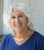 Sara Rosembaum has medium-length wavy white hair. She is wearing a blue dress.