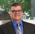 Ricardo Azziz has short dark hair and glasses. He is standing by a window with some greenery in the background.