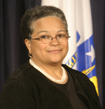 JudyAnn Bigby has very short coily gray hair. She is wearing a beaded necklace, glasses, and a black blouse with a black cardigan.