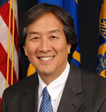 Howard Koh has short black/gray hair and is wearing a suit. He is sitting in front of some flags.