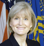 Mary Wakefield has short blonde/gray hair. She is sitting in front of some flags.