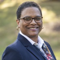 Felicia Collins is standing outside wearing a blue blazer, white shirt, and patterned scarf. She has cropped coily hair and glasses.
