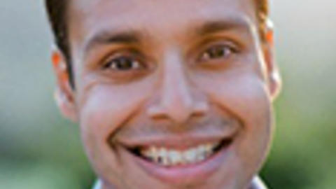 Joshua Budhu has short black hair. He is standing outside and wearing a blue shirt.
