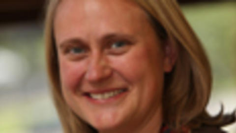 Anne Newland has medium length blonde hair. She is wearing a patterned blouse and blazer.