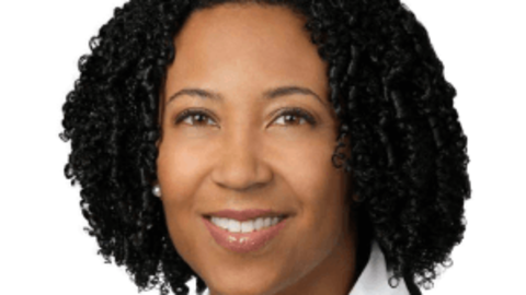 Dora Hughes is wearing a black blazer and white shirt. She has chin-length curly black hair.
