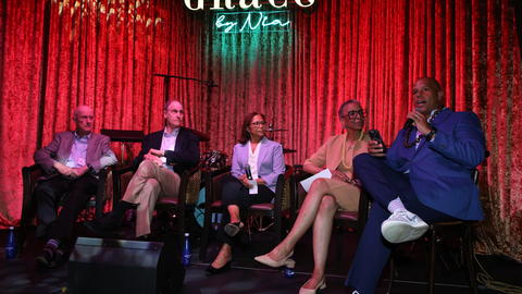 The fireside chat participants sit on a stage in front of a red curtain and underneath a neon Grace by Nia sign.