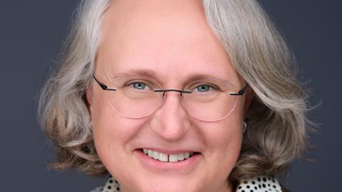 Anne Newland has medium length blonde/white hair. She is wearing a polka dot blouse and green blazer.