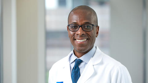 Jean Raphael is a bald man wearing glasses, a blue tie, and white doctor's coat.