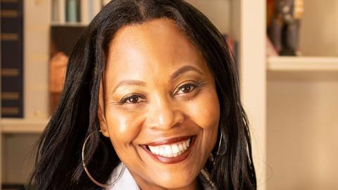 Christina Rosenthal has long black hair. She is wearing a white coat and sitting in front of a bookshelf.