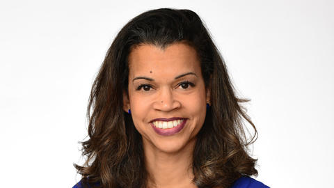 Shairi Turner has medium-length black hair. She is wearing a blue dress and is crossing her arms and smiling.
