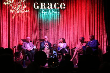 A group of people sit on a stage with a neon Grace By Nia sign above them. There is a red curtain in the background.