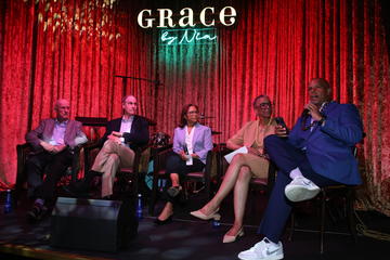 The fireside chat participants sit on a stage in front of a red curtain and underneath a neon Grace by Nia sign.