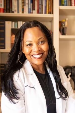 Christina Rosenthal has long black hair. She is wearing a white coat and sitting in front of a bookshelf.