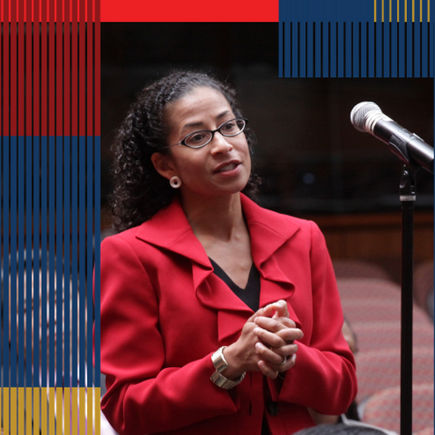 A woman in a red blazer speaks into a microphone next to her.