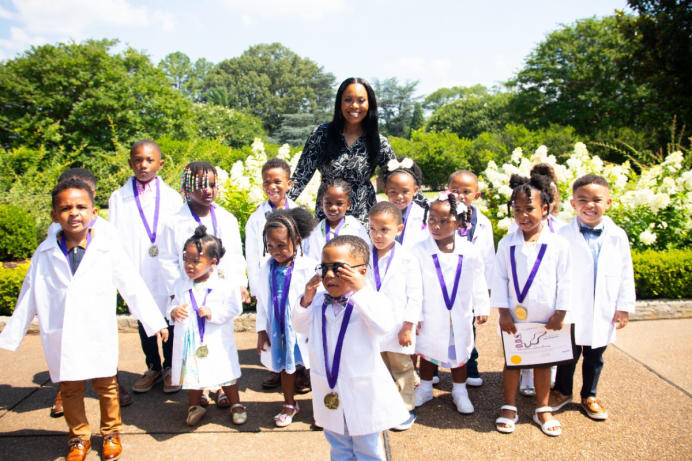 Rosenthal stands amongst a group of children all dressed in white coats. They are outside in front of a bunch of greenery.