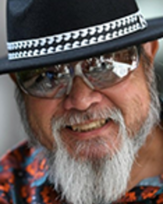 Ricardo Custodio is wearing a beaded black hat, silver sunglasses, and a printed shirt. He has a gray beard and moustache.