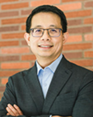 Gilbert Gee has short black hair and glasses. He is standing in front of a brick wall with his arms crossed.