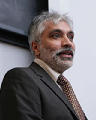Sanjay Gulati has gray hair and facial hair. He is wearing a brown suit and looking off to the side.