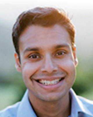 Joshua Budhu has short black hair. He is standing outside and wearing a blue shirt.