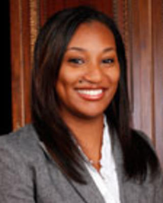 Kamillah Wood has long straight black hair. She is wearing a white shirt and gray blazer.