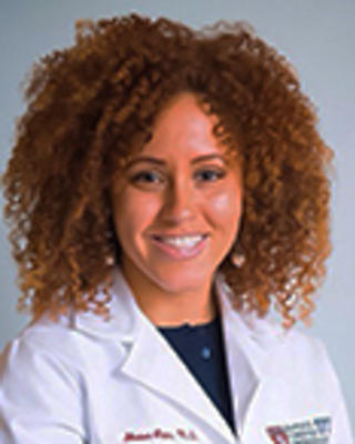 Shaina Lipa has medium length curly brown hair. She is wearing a white coat.