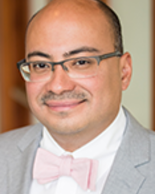 Lenny Lopez is a bald man with glasses and a moustache. He is wearing a white shirt, pink tie, and light gray blazer.