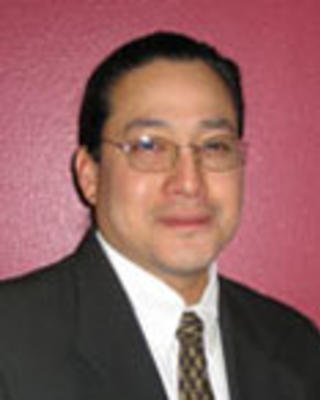 Lyle Ignace has black hair tied up and glasses. He is wearing a suit.