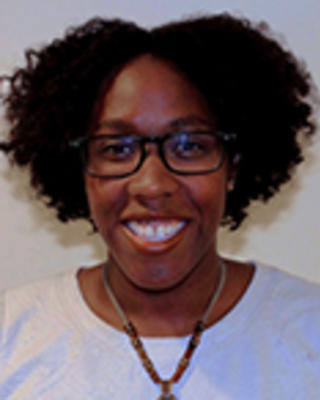 Paula Magee has short curly black hair and glasses. She is wearing a white shirt and pendant necklace.