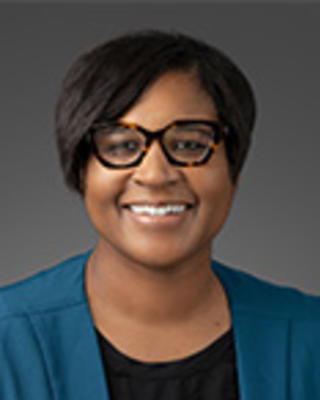 Morgan Medlock has short black hair and glasses. She is wearing a black shirt and blue jacket.