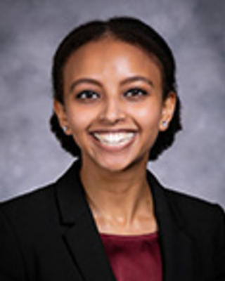 Meron Teklu has short coily hair tied back and is wearing a maroon blouse with a black blazer.
