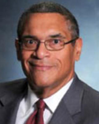 Woodrow Myers has very short black hair and glasses. He is wearing a suit.