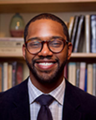Phillip Murray has very short black hair and facial hair. He is posed in front of a bookshelf and wearing a suit.