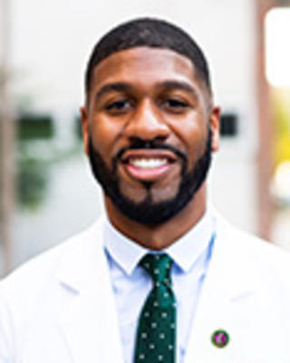Raymond Dawkins has short black hair and facial hair. He is wearing a white coat and green patterned tie.