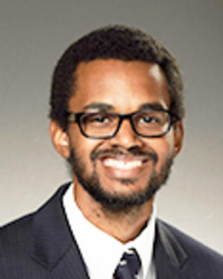 Alexander Rodgers has short coily black hair, facial hair, and glasses. He is wearing a suit.