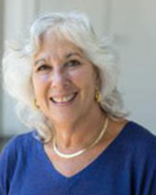 Sara Rosembaum has medium-length wavy white hair. She is wearing a blue dress.