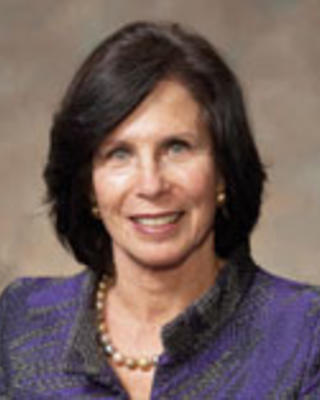 Gail Wilensky has short dark hair and is wearing a purple patterned jacket and pearl necklace.