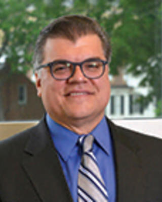 Ricardo Azziz has short dark hair and glasses. He is standing by a window with some greenery in the background.