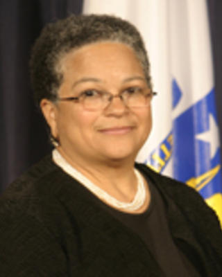 JudyAnn Bigby has very short coily gray hair. She is wearing a beaded necklace, glasses, and a black blouse with a black cardigan.