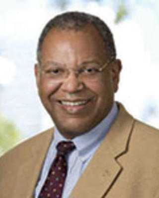 Otis Brawley has very short coily gray hair and glasses. He is wearing a beige suit.