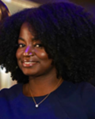 Magdala Chery has short curly black hair. She is wearing a blue dress and necklace.