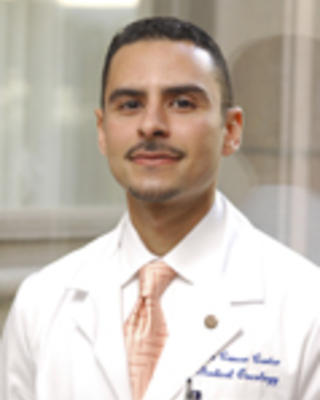 Aymen Elfiky is wearing a white coat, white shirt, and peach tie. He has short brown hair and a moustache.