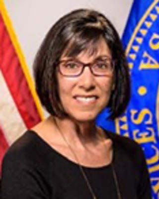Judith Steinberg has a bob and glasses. She is posed in front of an American flag and US seal.