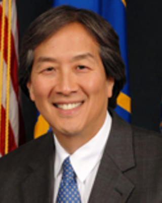 Howard Koh has short black/gray hair and is wearing a suit. He is sitting in front of some flags.