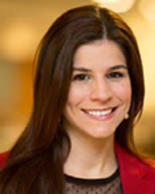 Maria Portela has straight long brown hair. She is wearing a black blouse and red blazer.