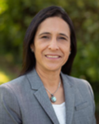 Nicole Stern has long straight black hair. She is standing outside and wearing a pendant necklace, white shirt, and gray blazer.